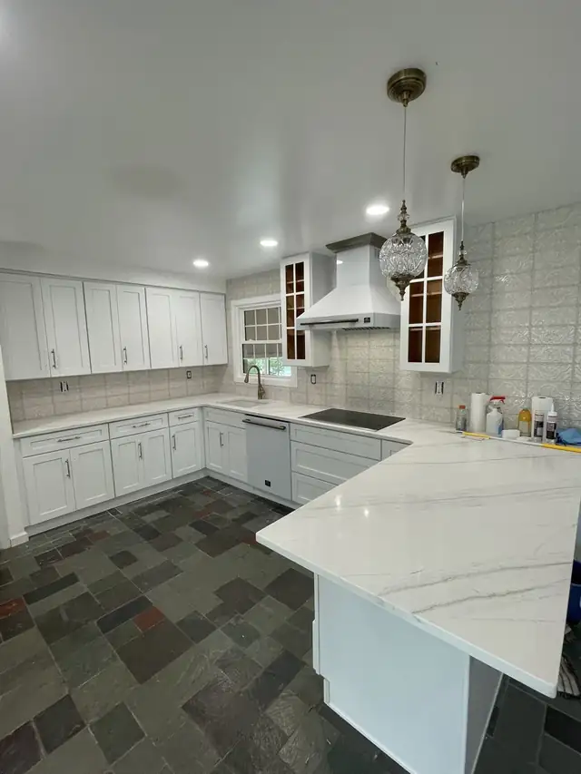 White shaker kitchen with veined quartz counters, white range hood, glass-front cabinets, crystal pendants and slate tile floor